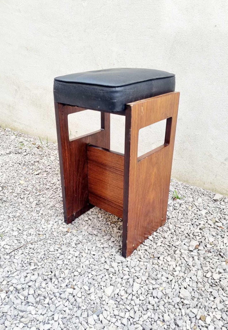Puede incluir: Taburete de madera vintage con asiento acolchado negro. El taburete tiene un asiento rectangular y un dise&ntilde;o de marco abierto con detalles recortados. La madera tiene un tono marr&oacute;n rojizo c&aacute;lido, sobre grava.