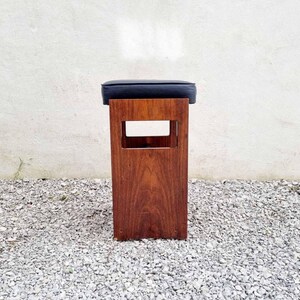 Puede incluir: Un taburete de madera con un asiento de coj&iacute;n azul oscuro. El taburete tiene un marco rectangular con un dise&ntilde;o recortado. La madera tiene un tono marr&oacute;n rojizo c&aacute;lido. El taburete est&aacute; colocado sobre una superficie de grava.