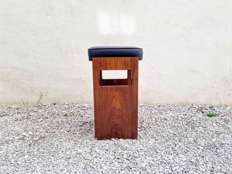 Puede incluir: Un taburete de madera con un asiento de coj&iacute;n azul oscuro. El taburete tiene un marco rectangular con un dise&ntilde;o recortado. La madera tiene un tono marr&oacute;n rojizo c&aacute;lido. El taburete est&aacute; colocado sobre una superficie de grava.