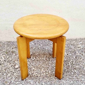 May include: A round wooden side table with a light brown finish and four legs. The table is sitting on a gravel surface.