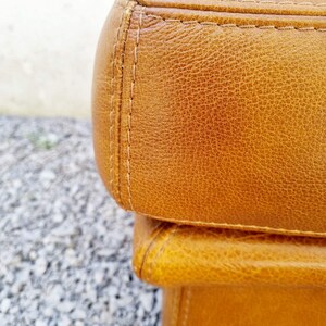 May include: Close-up of a section of a brown leather ottoman. The leather has a textured appearance with visible grain and subtle color variations. The edges are neatly stitched with a contrasting thread, highlighting the craftsmanship.