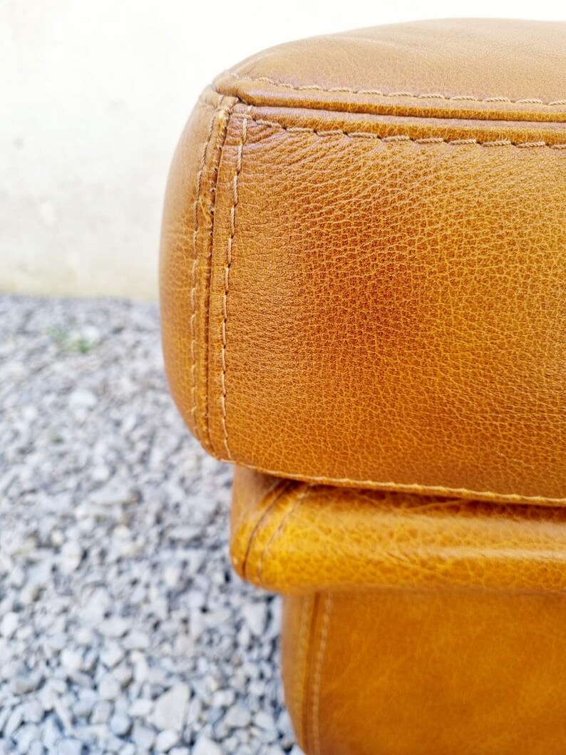 May include: Close-up of a section of a brown leather ottoman. The leather has a textured appearance with visible grain and subtle color variations. The edges are neatly stitched with a contrasting thread, highlighting the craftsmanship.