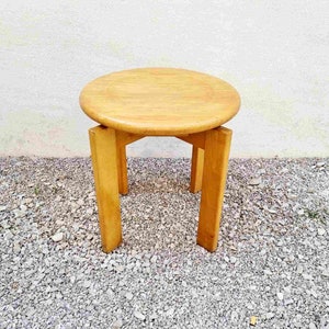 May include: A round wooden side table with a light brown finish and four legs. The table is sitting on a gravel surface.