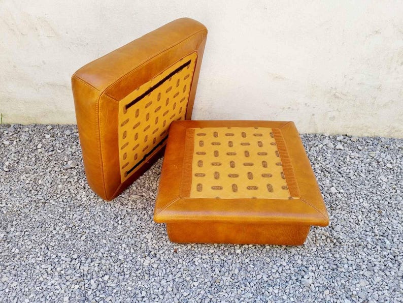 May include: Two-piece set of vintage furniture, featuring a square ottoman and a matching backrest. Both pieces are upholstered in tan leather with a patterned fabric insert. The ottoman measures approximately 18 inches square.