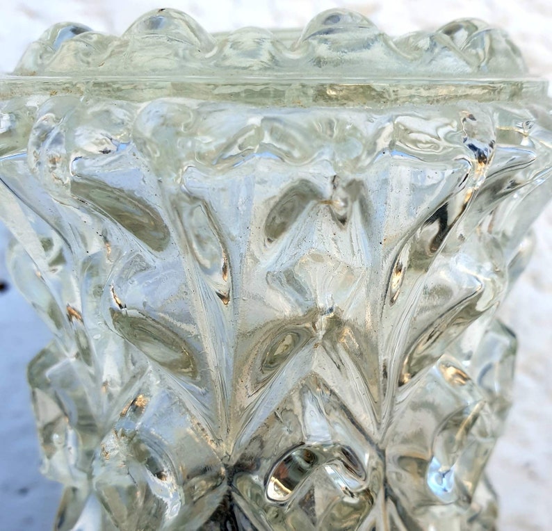 May include: A clear glass vase with a textured, geometric design. The vase has a wide mouth and a narrow base.