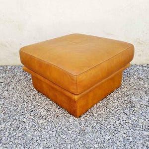 May include: A square, light brown leather ottoman. The ottoman has a two-tiered design with a padded top and a lower base. The piece is set on a bed of small, gray gravel.