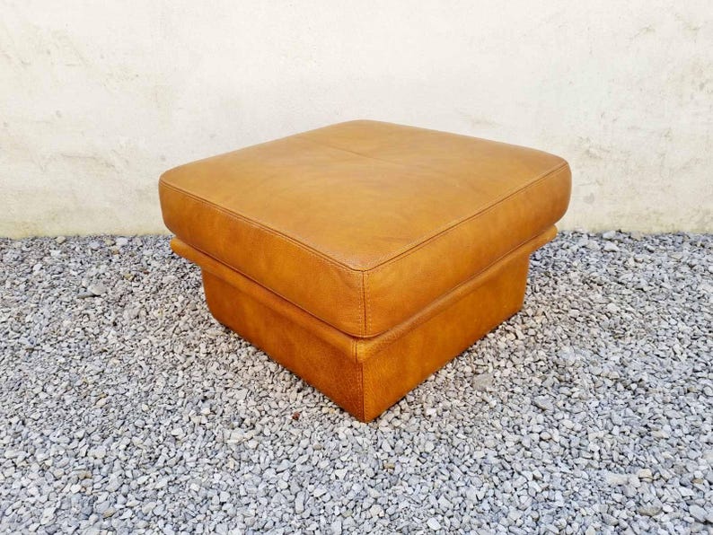 May include: A square, light brown leather ottoman. The ottoman has a two-tiered design with a padded top and a lower base. The piece is set on a bed of small, gray gravel.