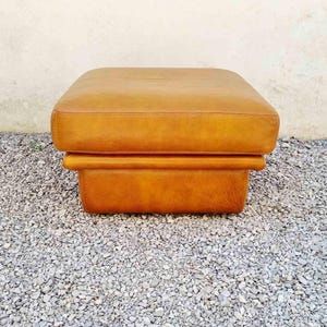 May include: A square, tan leather ottoman. The ottoman has a padded top and a lower section that forms a base. The leather has a slightly distressed appearance. The ottoman is sitting on a bed of small gray stones.