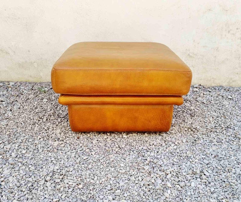May include: A square, tan leather ottoman. The ottoman has a padded top and a lower section that forms a base. The leather has a slightly distressed appearance. The ottoman is sitting on a bed of small gray stones.
