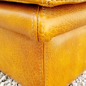 May include: Close-up of a section of a mustard-colored leather object. The leather has a textured, cracked appearance with visible stitching along the seams. The object appears to be a piece of furniture.