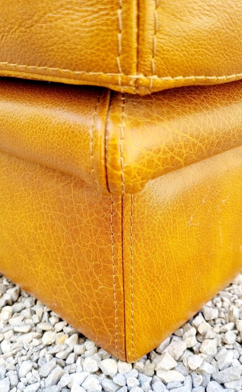May include: Close-up of a section of a mustard-colored leather object. The leather has a textured, cracked appearance with visible stitching along the seams. The object appears to be a piece of furniture.