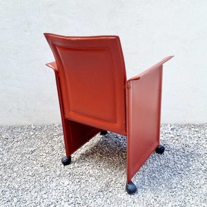 May include: A red leather office chair with wheels. The chair has a high back and arms. The chair is on a gravel surface.