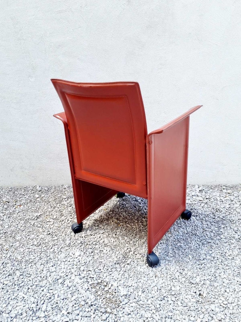 May include: A red leather office chair with wheels. The chair has a high back and arms. The chair is on a gravel surface.