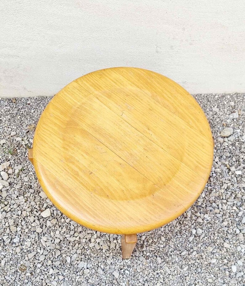 May include: A round wooden stool with a light brown finish. The stool has three legs and a smooth, slightly worn surface.