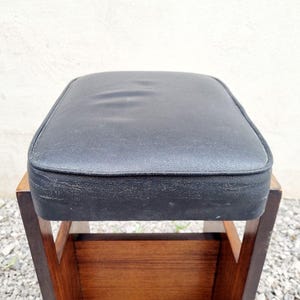 Puede incluir: Un taburete vintage con un asiento acolchado negro y cuadrado. El asiento est&aacute; enmarcado por un borde oscuro y se asienta sobre un marco de madera con un estante inferior. La madera tiene un tono marr&oacute;n c&aacute;lido y el taburete parece usado.