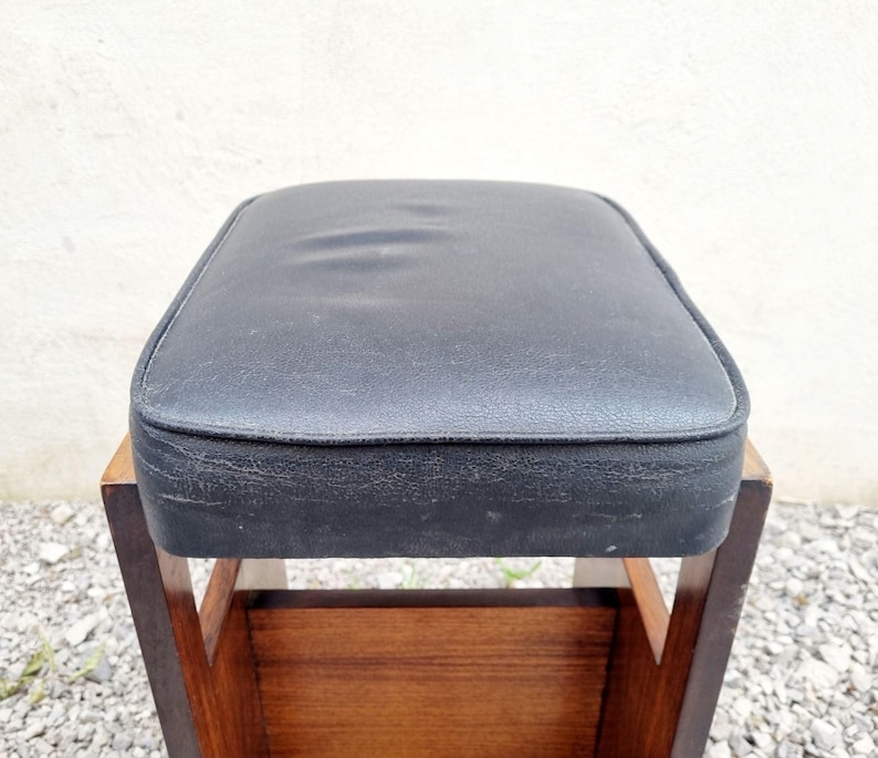 Puede incluir: Un taburete vintage con un asiento acolchado negro y cuadrado. El asiento est&aacute; enmarcado por un borde oscuro y se asienta sobre un marco de madera con un estante inferior. La madera tiene un tono marr&oacute;n c&aacute;lido y el taburete parece usado.