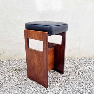 Puede incluir: Taburete moderno de mediados de siglo con asiento acolchado azul oscuro. El taburete tiene un marco rectangular de madera con un acabado marr&oacute;n c&aacute;lido. El dise&ntilde;o incluye detalles recortados en los laterales. El taburete est&aacute; sobre una superficie de grava. Altura aproximada 45 cm.