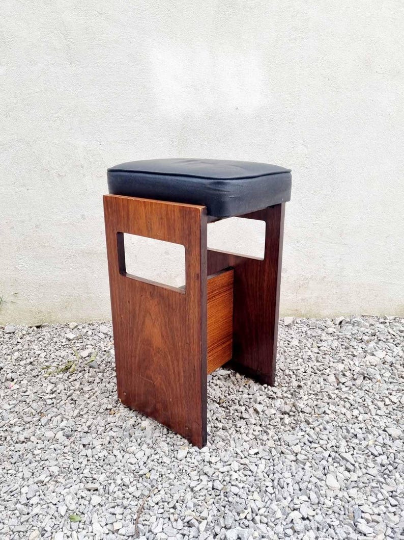 Puede incluir: Taburete moderno de mediados de siglo con asiento acolchado azul oscuro. El taburete tiene un marco rectangular de madera con un acabado marr&oacute;n c&aacute;lido. El dise&ntilde;o incluye detalles recortados en los laterales. El taburete est&aacute; sobre una superficie de grava. Altura aproximada 45 cm.