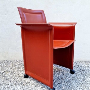 May include: A red leather chair with a square back and a square seat. The chair has four wheels and is on a gravel surface.