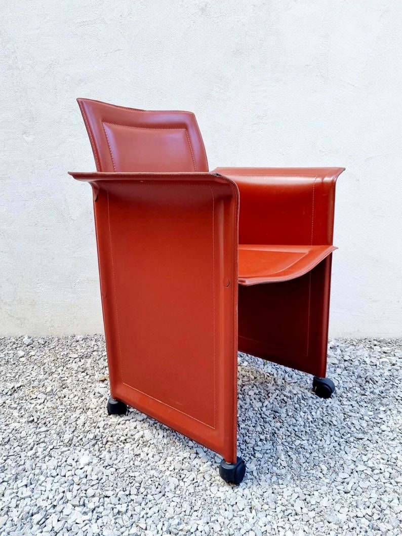 May include: A red leather chair with a square back and a square seat. The chair has four wheels and is on a gravel surface.