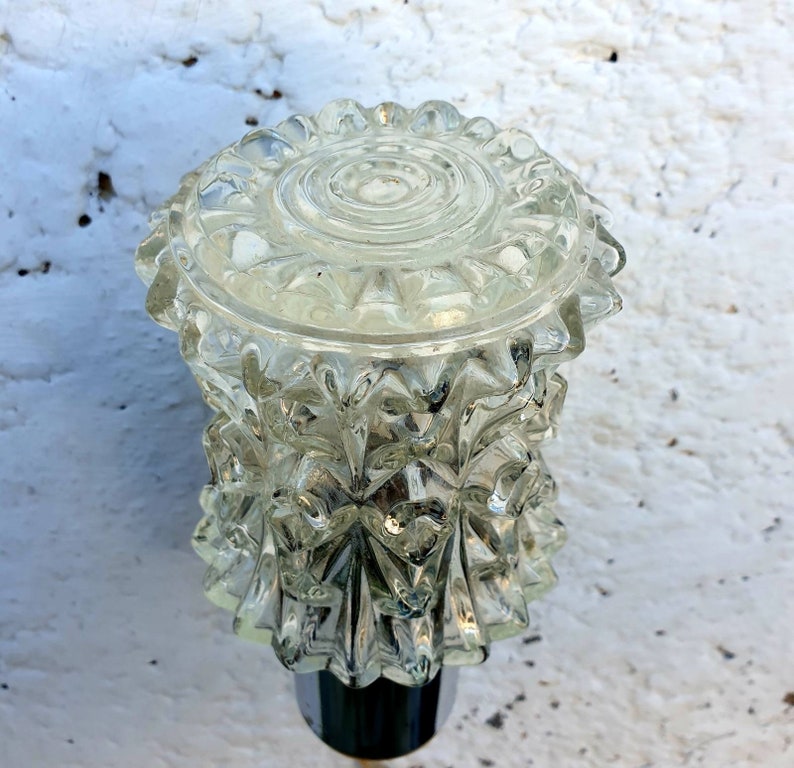 May include: A clear glass lampshade with a textured, faceted design. The lampshade has a round top and a pointed bottom.