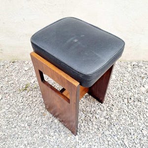 Puede incluir: Un peque&ntilde;o taburete vintage con un asiento rectangular de coj&iacute;n negro. El marco de madera es de color marr&oacute;n c&aacute;lido, con un dise&ntilde;o recortado en los laterales. El taburete mide probablemente 46 cm de alto.