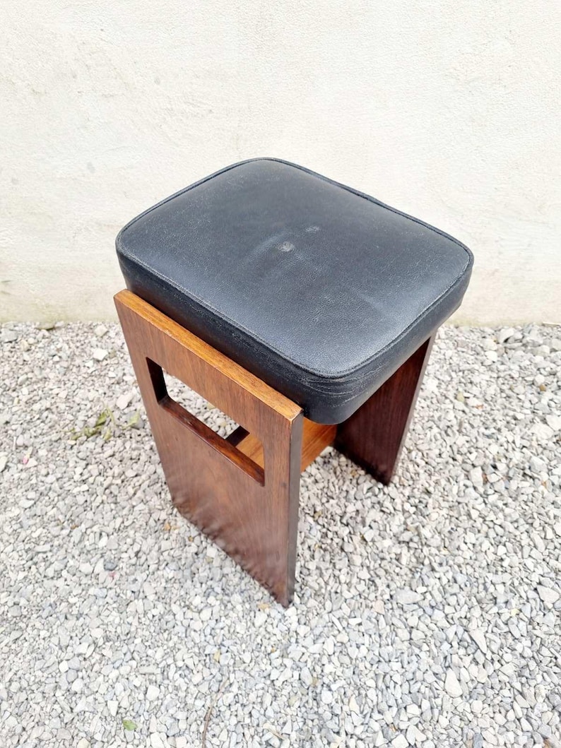 Puede incluir: Un peque&ntilde;o taburete vintage con un asiento rectangular de coj&iacute;n negro. El marco de madera es de color marr&oacute;n c&aacute;lido, con un dise&ntilde;o recortado en los laterales. El taburete mide probablemente 46 cm de alto.