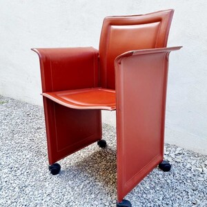 May include: A red leather chair with a square back and four wheels. The chair is on a gravel surface.