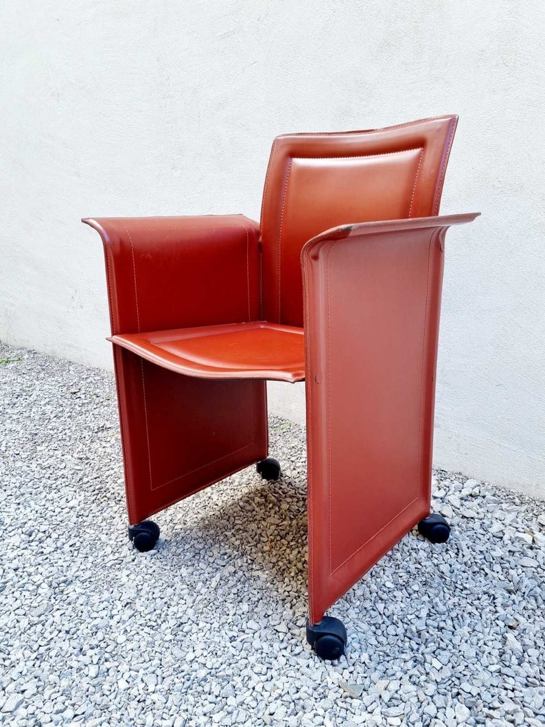 May include: A red leather chair with a square back and four wheels. The chair is on a gravel surface.