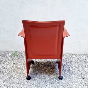 May include: A red leather chair with a square back and a metal frame. The chair has four wheels and is sitting on a gravel surface.