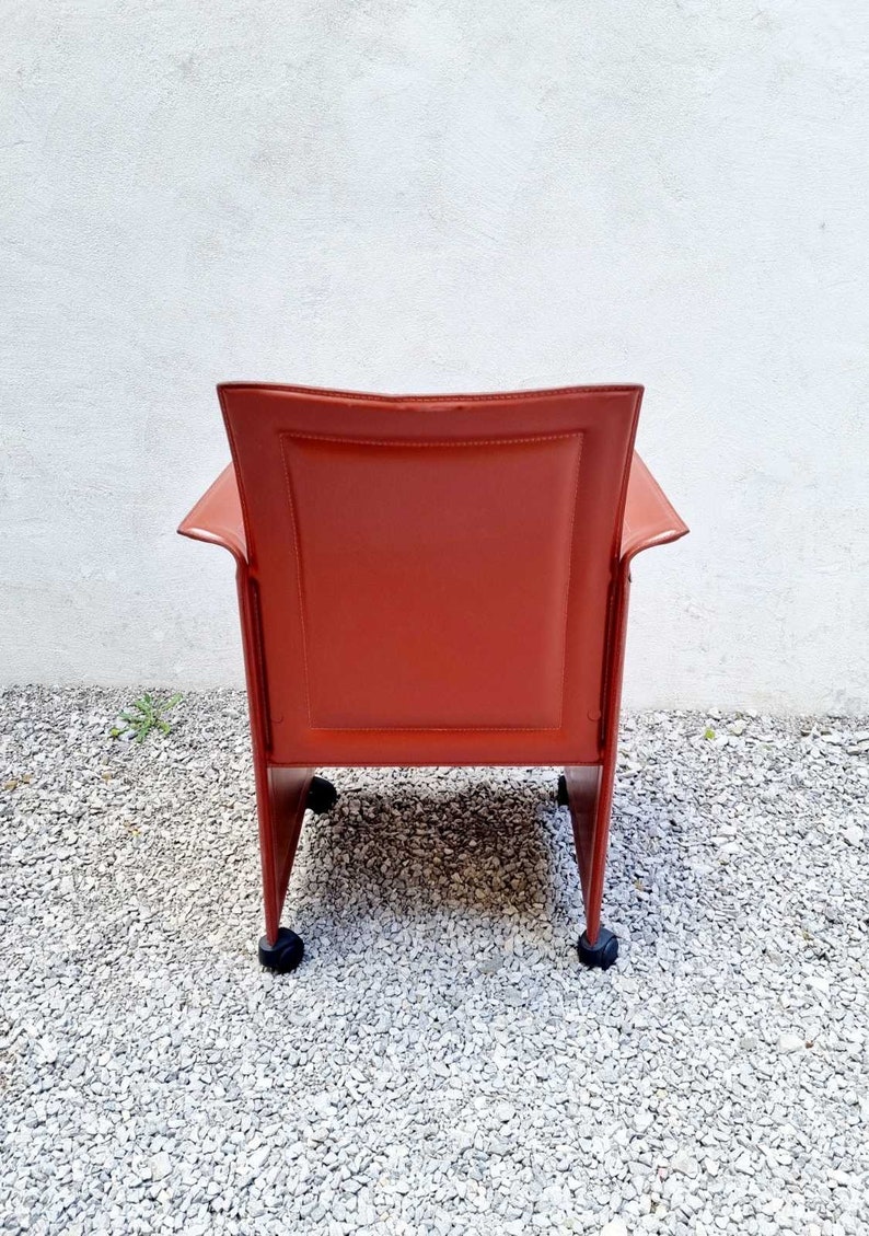 May include: A red leather chair with a square back and a metal frame. The chair has four wheels and is sitting on a gravel surface.