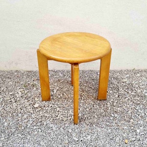 May include: A round wooden side table with a light brown finish and three legs. The table is sitting on a bed of small gray stones.