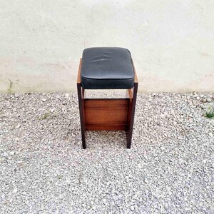 Puede incluir: Un taburete de madera vintage con un asiento acolchado negro. El taburete tiene un asiento rectangular y un estante inferior. El marco es de madera oscura. El taburete est&aacute; sobre una superficie de grava.