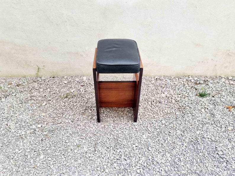Puede incluir: Un taburete de madera vintage con un asiento acolchado negro. El taburete tiene un asiento rectangular y un estante inferior. El marco es de madera oscura. El taburete est&aacute; sobre una superficie de grava.