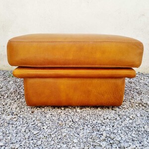 May include: A rectangular, two-tiered ottoman in a warm, golden-brown leather. The top cushion is slightly larger than the base, creating a layered effect. The ottoman is placed on a bed of small, gray gravel.