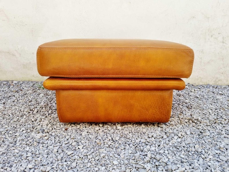 May include: A rectangular, two-tiered ottoman in a warm, golden-brown leather. The top cushion is slightly larger than the base, creating a layered effect. The ottoman is placed on a bed of small, gray gravel.
