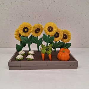 Op de afbeelding: Een miniatuur tuinscène met een houten plantenbak gevuld met kleine plastic zonnebloemen, bloemkool, wortels en pompoenen.