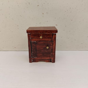 May include: A miniature wooden nightstand with a dark reddish-brown finish. It features a small drawer with a brass knob and a cabinet door with a matching knob. The nightstand is a small piece of furniture, likely for a dollhouse or miniature setting.