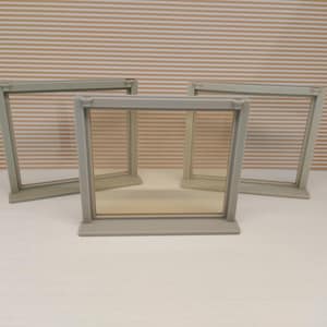May include: Three gray rectangular frames with clear glass panels. The frames are arranged on a white surface against a backdrop of horizontal beige and white stripes. The frames are likely for displaying photos or artwork.