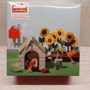 May include: A Lundby dollhouse accessory set featuring a dog house, a dog, a rake, a watering can, a bowl, a pumpkin, carrots, a cabbage, sunflowers, and a mailbox. The set is on a green lawn with a white picket fence.