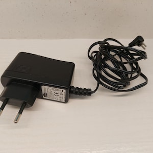 May include: A black power adapter with a two-prong plug and a coiled black cord. The adapter has a rectangular body and a label with technical information. The cord is neatly coiled, ready for use.