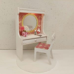 Puede incluir: Conjunto de tocador blanco con un respaldo estampado floral rosa, un espejo redondo, un cajón y una silla a juego. El tocador está sobre una base blanca.
