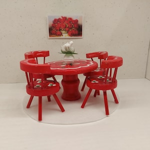 May include: Miniature red wooden dining set with a round table and four chairs. The table and chairs are decorated with white floral accents. A small vase with white flowers sits on the table. A framed picture of red flowers hangs on the wall.