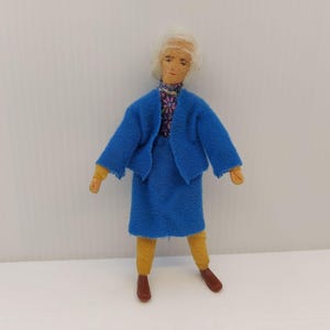 May include: A handmade doll dressed in a blue skirt suit with a floral patterned scarf. The doll has blonde hair, yellow arms and legs, and brown shoes. The doll is standing against a white background.