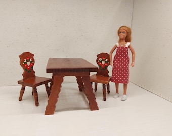 May include: Miniature wooden table and chairs set. The table has a dark brown finish with a unique leg design. Two chairs with decorative floral accents flank the table. A doll in a red polka dot dress stands nearby.