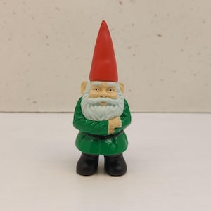 May include: A small garden gnome figurine. The gnome has a red pointed hat, a white beard, and is wearing a green jacket and black boots. The gnome is standing with its arms crossed.