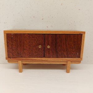 May include: A small wooden cabinet with two doors and brass knobs. The cabinet has a light brown top and bottom with a darker brown wood for the doors.