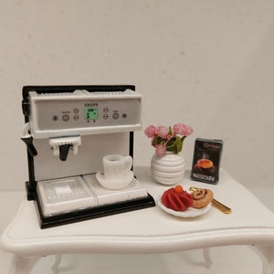 Puede incluir: Una máquina de espresso blanca en miniatura con una base negra, una taza y un platillo blancos, un arreglo de flores rosas en un jarrón blanco, una caja de cápsulas de café Nescafe Espresso, un pastel rojo con una cobertura amarilla y un pastel marrón en un plato blanco.