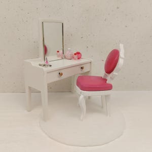 Op de afbeelding: Een miniatuur witte kaptafelset met een spiegel, een lade en decoratieve flesjes. Een bijpassende stoel met een roze zitting en rugleuning staat naast de kaptafel. De set staat op een wit tapijt.