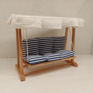 May include: A miniature wooden swing with a white canopy and a blue and white striped cushion. The swing is suspended by ropes and has a light brown wooden frame. The canopy is made of white fabric.